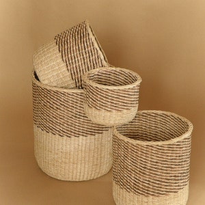 May include: A set of three cylindrical woven baskets featuring a two-tone design. Each basket is constructed with light beige natural fibers at the base and a contrasting dark brown woven pattern around the upper section, creating a distinct visual divide.