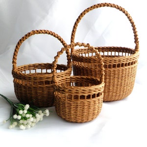 May include: Three woven brown baskets of different sizes, each with a rounded handle. The baskets are made of a light brown material and have a textured appearance. A sprig of white flowers is in the lower left corner.