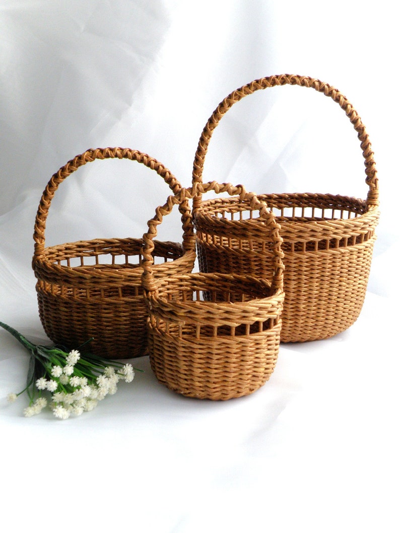 May include: Three woven brown baskets of different sizes, each with a rounded handle. The baskets are made of a light brown material and have a textured appearance. A sprig of white flowers is in the lower left corner.