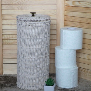 Puede incluir: Porta rollo de papel higi&eacute;nico de mimbre blanco con un pomo de lat&oacute;n en la parte superior. Tres rollos de papel higi&eacute;nico blanco con un dise&ntilde;o floral verde est&aacute;n apilados junto al soporte.