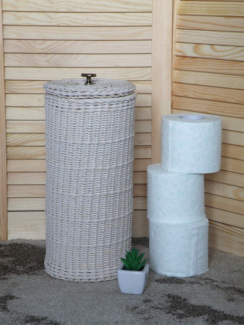 Puede incluir: Porta rollo de papel higi&eacute;nico de mimbre blanco con un pomo de lat&oacute;n en la parte superior. Tres rollos de papel higi&eacute;nico blanco con un dise&ntilde;o floral verde est&aacute;n apilados junto al soporte.