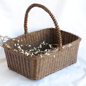 May include: A rectangular brown wicker basket with a braided handle. The basket contains white floral stems. The basket is made of woven material and is suitable for storage or decoration.