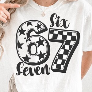 May include: White t-shirt with the numbers "67" in a black, bold font. The number "6" is filled with stars, and the number "7" has a checkered pattern. The words "Six" and "Seven" are written above and below the numbers.