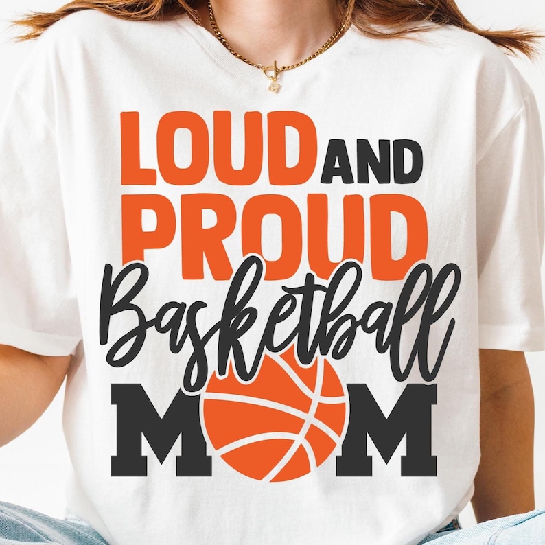 K&ouml;nnte beinhalten: Wei&szlig;es T-Shirt mit dem Text "LOUD AND PROUD Basketball MOM" in Orange und Schwarz. Eine orangefarbene Basketballgrafik ist unter dem Text zentriert. Das Shirt hat einen Rundhalsausschnitt.