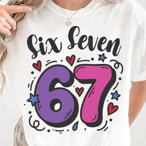 May include: White t-shirt featuring the words "Six Seven" in black script above a large, colourful "67" in purple and pink. The design includes red hearts, blue stars, and decorative swirls.
