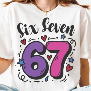 May include: White t-shirt featuring the words "Six Seven" in black script. The numbers "67" are in purple and pink, outlined in black, with hearts and stars. A gold necklace is visible.