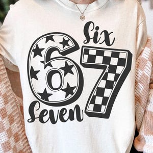 May include: Cream-coloured sweatshirt with a black graphic design. The design features the word "Six" above the numbers "67" and the word "Seven" in a cursive font. The number "6" is a circle filled with stars, and the "7" has a chequered pattern.