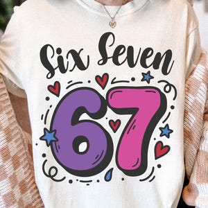 May include: A cream-coloured t-shirt featuring the words "Six Seven" in black script above a large, outlined number "67". The "6" is purple, and the "7" is pink. Small hearts, stars, and dots surround the numbers.