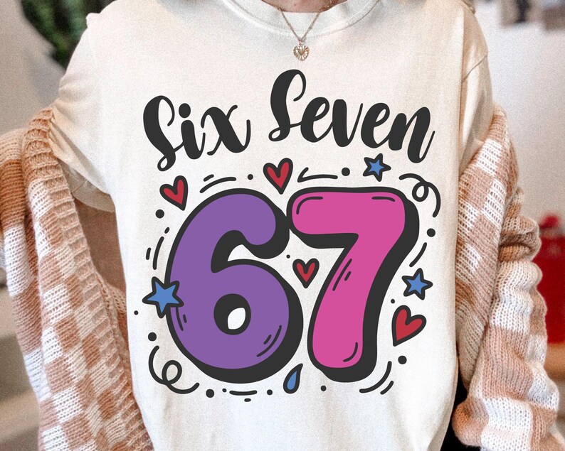 May include: A cream-coloured t-shirt featuring the words "Six Seven" in black script above a large, outlined number "67". The "6" is purple, and the "7" is pink. Small hearts, stars, and dots surround the numbers.