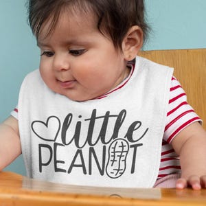 Puede incluir: Un babero blanco para beb&eacute; con el texto "little peanut" en letras negras y un peque&ntilde;o coraz&oacute;n sobre la palabra "little". Un gr&aacute;fico de cacahuete est&aacute; al lado de la palabra "peanut". Un beb&eacute; lleva el babero y mira hacia abajo.
