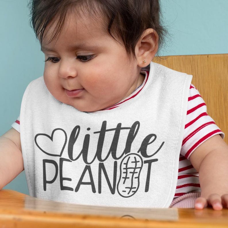 Puede incluir: Un babero blanco para beb&eacute; con el texto "little peanut" en letras negras y un peque&ntilde;o coraz&oacute;n sobre la palabra "little". Un gr&aacute;fico de cacahuete est&aacute; al lado de la palabra "peanut". Un beb&eacute; lleva el babero y mira hacia abajo.