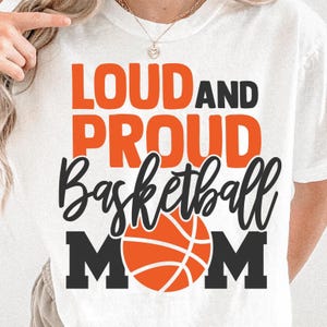 K&ouml;nnte beinhalten: Wei&szlig;es T-Shirt mit dem Text "LOUD AND PROUD Basketball MOM" in Orange und Schwarz. Das Wort "Basketball" ist in einer Schreibschrift geschrieben, und eine Basketball-Grafik befindet sich in der Mitte.