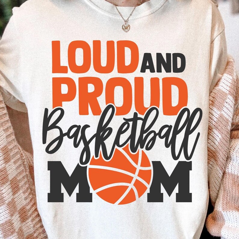 K&ouml;nnte beinhalten: Cremefarbenes T-Shirt mit den Worten "LOUD AND PROUD Basketball MOM" in Orange und Schwarz. Eine Basketball-Grafik ist unter dem Text zentriert. Das Shirt ist ein l&auml;ssiger Stil.