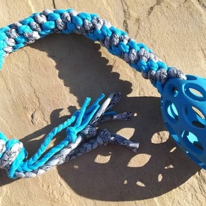 May include: A dog toy featuring a blue and white braided rope leading to a blue, oval-shaped ball. The braided section is made of blue, white, and grey fabric. The toy is designed for interactive play.