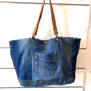 Peut inclure: Un sac fourre-tout en jean avec des anses en cuir marron. Le sac est doté d'une poche avant avec des rivets décoratifs et une fermeture boutonnée. La doublure intérieure présente un motif floral. Le sac est suspendu sur un fond blanc.