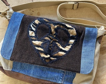 Pode incluir: Uma bolsa de mensageiro de lona marrom com um design de patchwork. A bolsa apresenta um grande sinal de paz feito de pele sintética com um padrão de zebra. A bolsa tem uma alça longa ajustável.