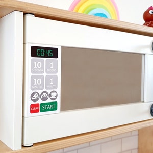 May include: A white toy microwave oven with a digital display showing 00:45. It has buttons for seconds, minutes, and food options. The handle is dark gray. The microwave is set in a wooden shelf.