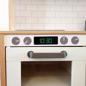 May include: A white toy kitchen with a wooden countertop and a white brick backsplash. The oven features a digital clock displaying 10:30 and grey knobs. A grey handle is on the oven door.