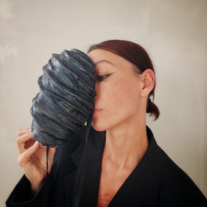 Pu&ograve; includere: Una persona che indossa un blazer nero tiene una maschera scultorea testurizzata nera davanti al viso. La maschera ha una forma organica unica e sembra essere fatta di carta o tessuto.
