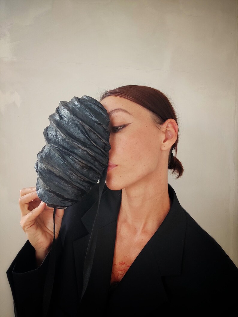 Pu&ograve; includere: Una persona che indossa un blazer nero tiene una maschera scultorea testurizzata nera davanti al viso. La maschera ha una forma organica unica e sembra essere fatta di carta o tessuto.