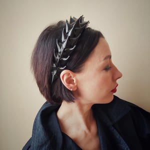 May include: A black headband with a spiky, leaf-like design. The headband is made of a shiny, textured material.