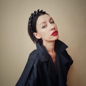 May include: A woman wearing a black jacket and a black headband with a geometric design. She has red lipstick on and is looking at the camera.