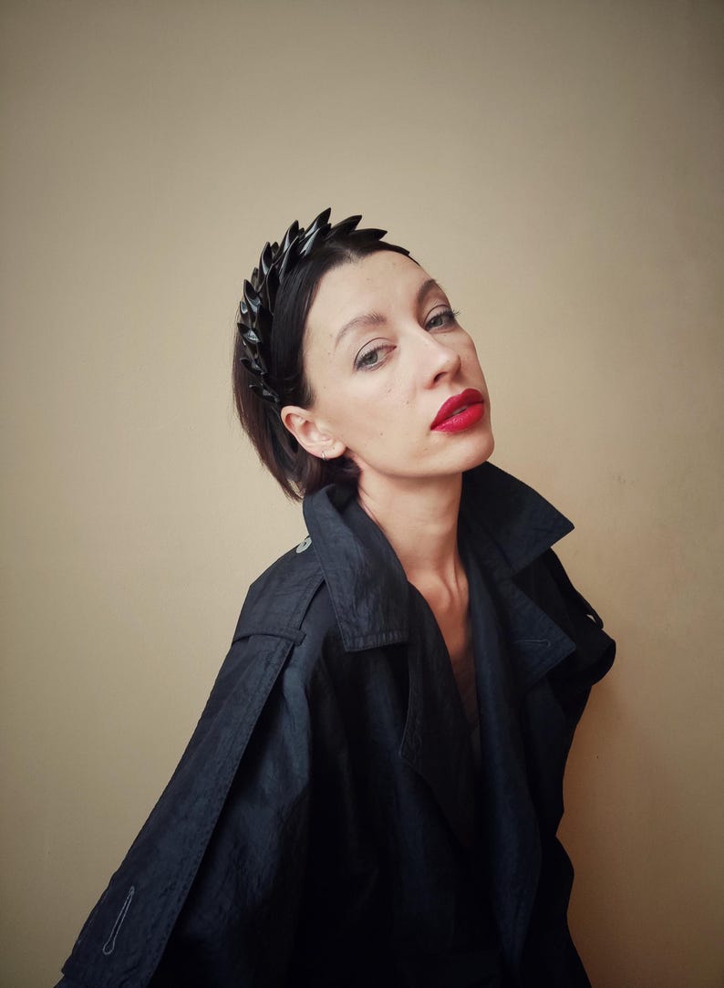 May include: A woman wearing a black jacket and a black headband with a geometric design. She has red lipstick on and is looking at the camera.