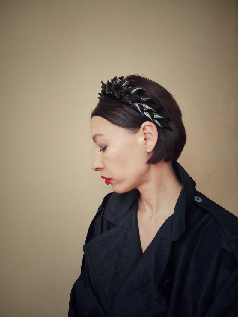 May include: A woman with short dark hair is wearing a black headband with a geometric design. She is wearing a black jacket and has red lipstick on.