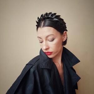 May include: A black headband with a jagged, leaf-like design. The headband is worn by a person with short, dark hair and red lipstick.