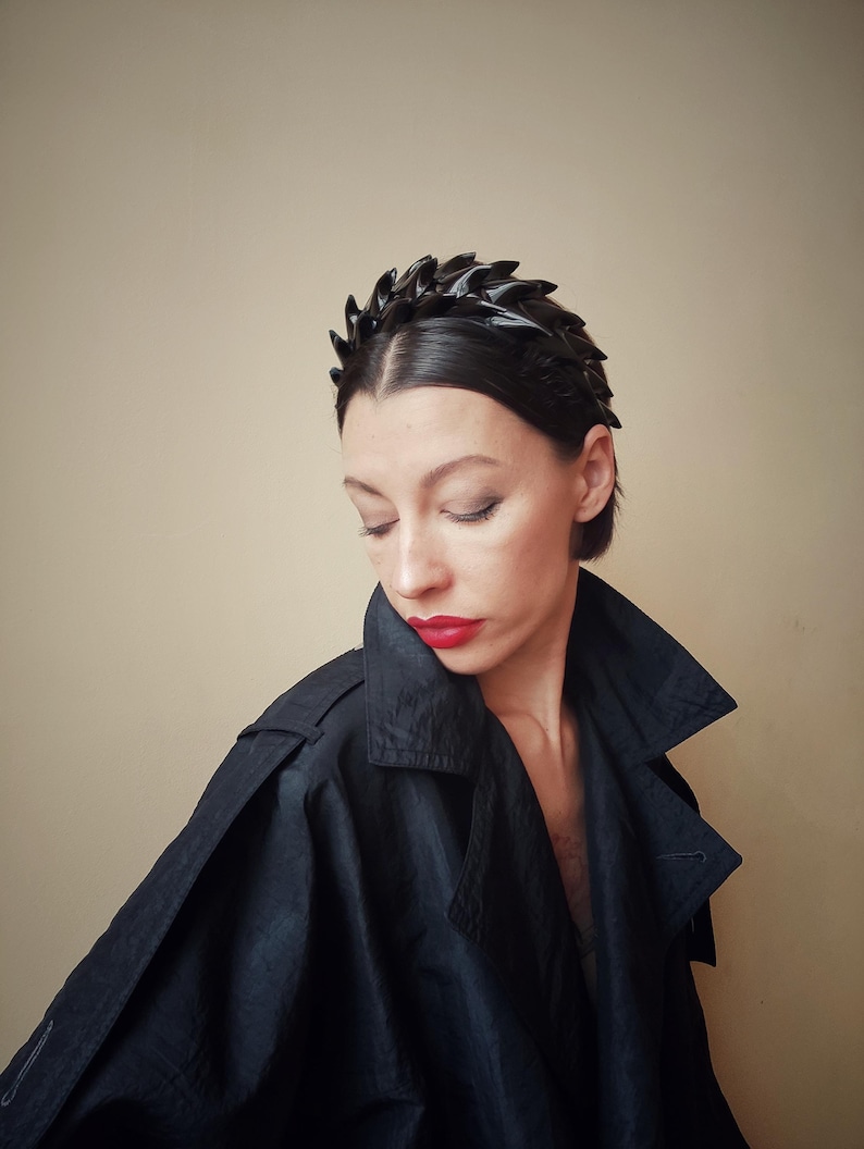 May include: A black headband with a jagged, leaf-like design. The headband is worn by a person with short, dark hair and red lipstick.