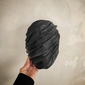 Pu&ograve; includere: Una maschera scultorea astratta nera con una superficie liscia e testurizzata. La maschera ha la forma di un viso e presenta una serie di linee curve che creano un senso di movimento.