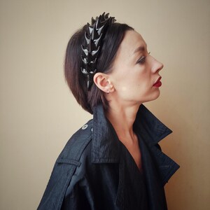 May include: A black headband with a row of sharp, pointed spikes. The headband is worn by a person with short, dark hair.