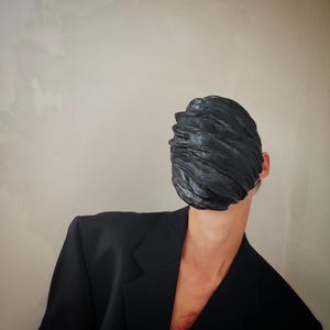 Pu&ograve; includere: Una persona che indossa un blazer nero con una maschera di stoffa nera che le copre il viso. La maschera &egrave; testurizzata e ha un aspetto stropicciato.