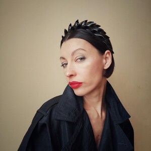 May include: A woman with short dark hair is wearing a black headband with a jagged edge. She is wearing a black jacket and has red lipstick on.