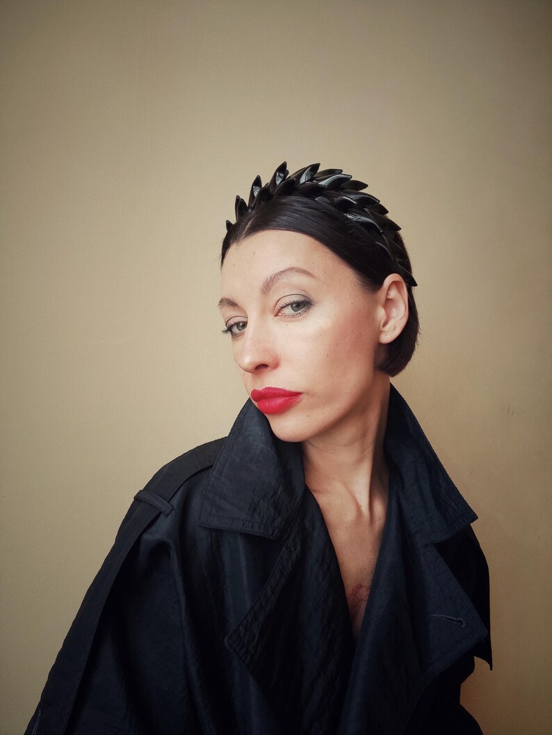 May include: A woman with short dark hair is wearing a black headband with a jagged edge. She is wearing a black jacket and has red lipstick on.