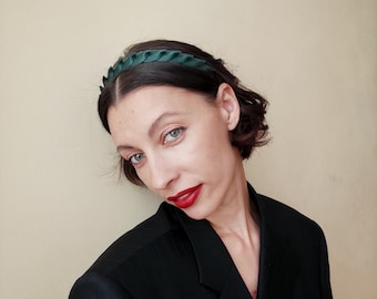 May include: A dark green satin headband with a braided design. The headband is worn on the head. The person is wearing a black blazer and has red lipstick.