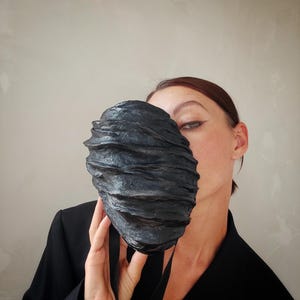 Pu&ograve; includere: Una maschera facciale nera con un design testurizzato e stratificato. La maschera &egrave; realizzata in un materiale morbido e flessibile e ha due lacci neri.