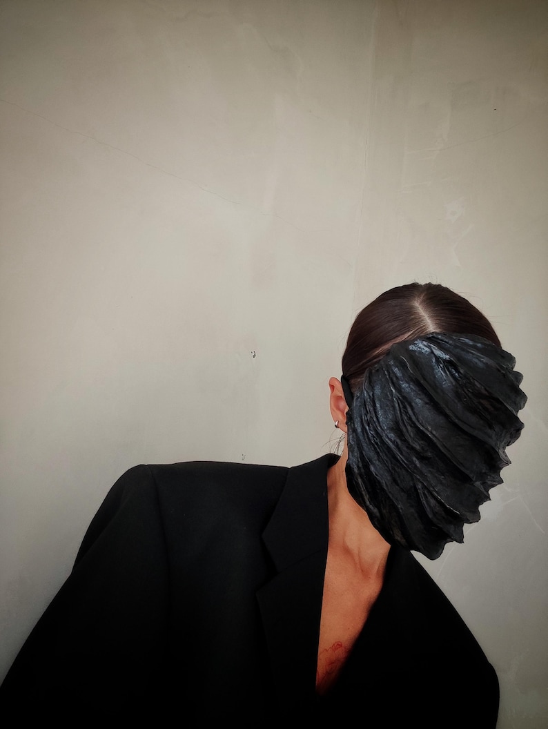 Pu&ograve; includere: Una persona che indossa un blazer nero e una maschera facciale nera con un design plissettato.