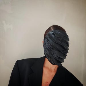 Pu&ograve; includere: Una persona che indossa un blazer nero con una maschera facciale nera testurizzata. La maschera copre l'intero viso della persona.