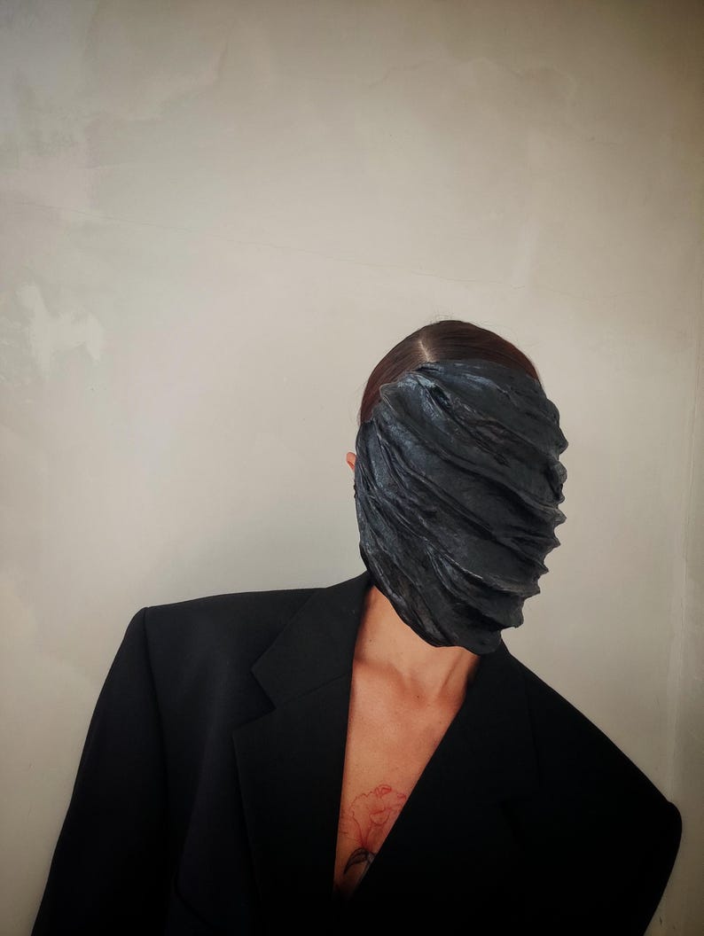 Pu&ograve; includere: Una persona che indossa un blazer nero con una maschera facciale nera testurizzata. La maschera copre l'intero viso della persona.