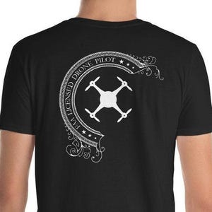 May include: A black short-sleeved t-shirt featuring a white graphic on the back. The design includes a central drone icon surrounded by a curved banner with the text FAA LICENSED DRONE PILOT and decorative flourishes at the ends of the banner.