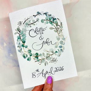 May include: A white wedding invitation featuring a watercolour eucalyptus wreath. The invitation includes the names "Colette & John" and the date "18th April 2026" in calligraphy. The wreath has gold detailing.