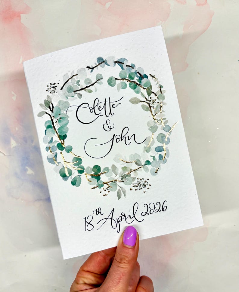 May include: A white wedding invitation featuring a watercolour eucalyptus wreath. The invitation includes the names "Colette & John" and the date "18th April 2026" in calligraphy. The wreath has gold detailing.
