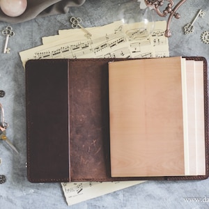 Peut inclure: Un journal en cuir marron avec un insert en papier blanc crème vierge. Le journal est ouvert et posé à plat sur une surface grise avec une feuille de musique en dessous. Divers objets décoratifs sont éparpillés autour du journal, notamment des clés, des engrenages et une plume.
