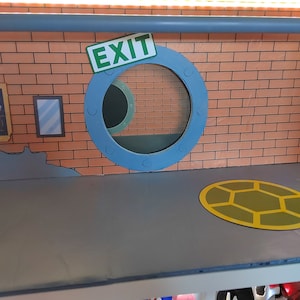 May include: A playset featuring a brick wall with a blue and white round window with the word "EXIT" on it. The playset has a gray floor with a yellow turtle shell design.