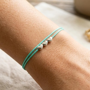 May include: A double-strand turquoise cord bracelet featuring three small, polished silver-toned heart charms centered on the cord. The bracelet is designed with a simple, minimalist aesthetic, showcasing the vibrant color of the cord and the metallic finish of the hearts.