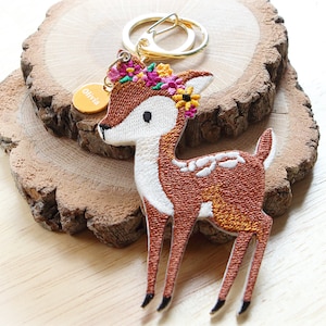 Könnte beinhalten: Ein brauner und weißer bestickter Hirsch-Schlüsselanhänger mit einer Blumenkrone und einem goldenen Anhänger mit der Aufschrift "Olivia".