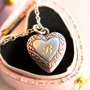 May include: A silver heart-shaped locket necklace with an ornate design and the letter "B" engraved in gold inside. The necklace is displayed in a pink velvet box.