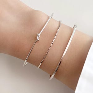 Puede incluir: Una colección de pulseras de plata en una muñeca. Las pulseras incluyen un diseño de nudo, una cadena de eslabones cuadrados y dos bandas simples. Las pulseras se muestran en la muñeca de una persona, con una camisa blanca visible en el fondo.