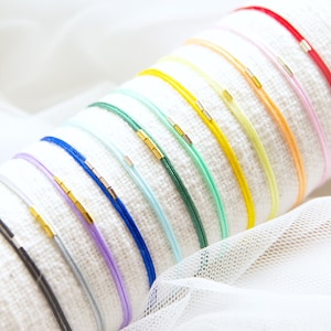 Puede incluir: Una colección de pulseras de cuerda coloridas con detalles metálicos dorados. Las pulseras se exhiben sobre una superficie blanca y texturizada, mostrando una variedad de colores vibrantes, incluyendo rojo, amarillo, azul y verde. Cada pulsera tiene un diseño simple y minimalista.